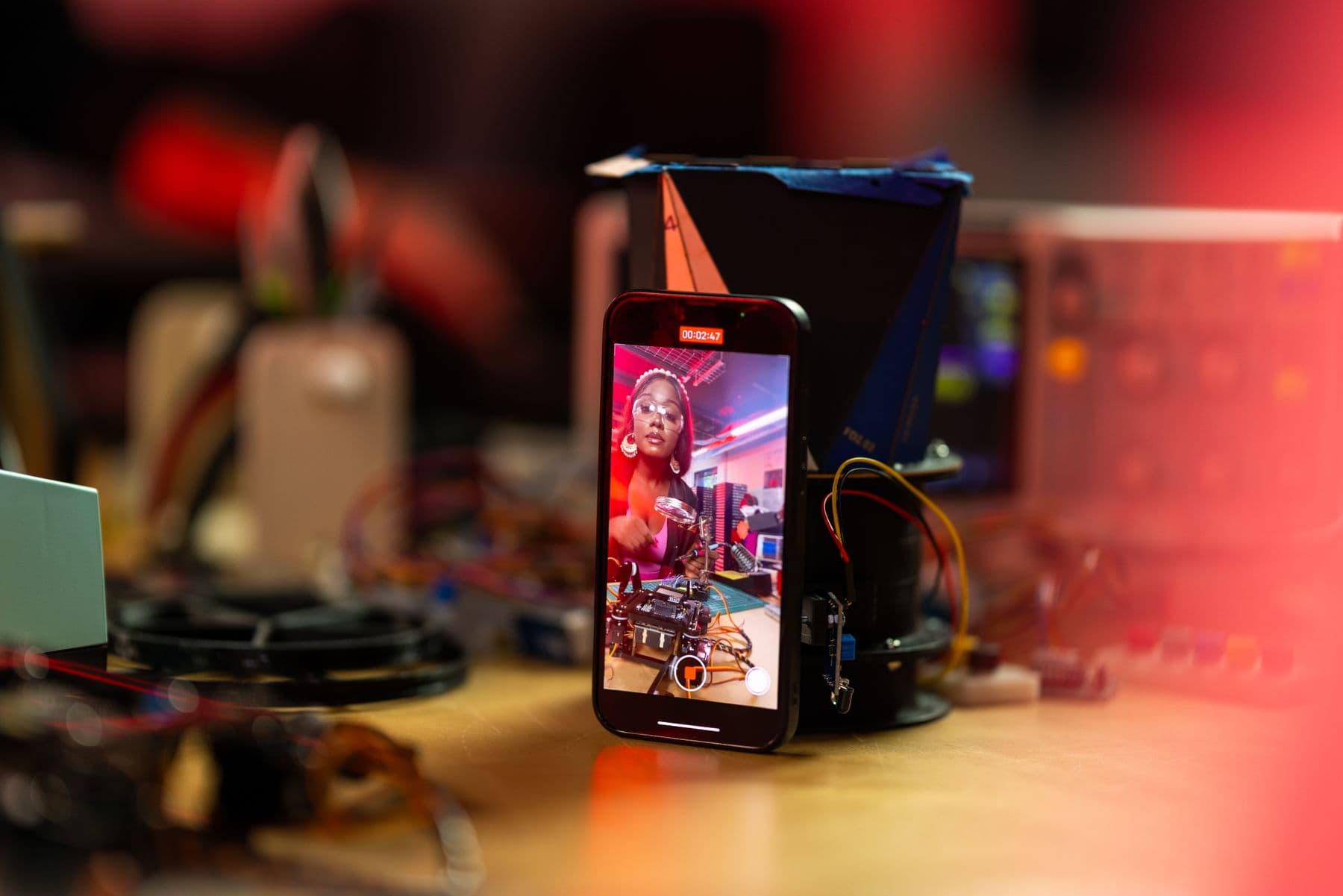 A phone recording a woman demonstrating electronics at a workbench.